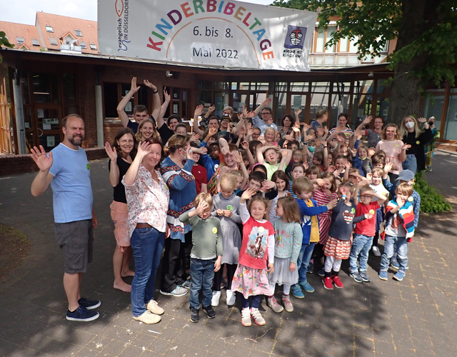Kinder und Erwachsene winken fröhlich draußen unter dem Kinderbibeltage-Banner.