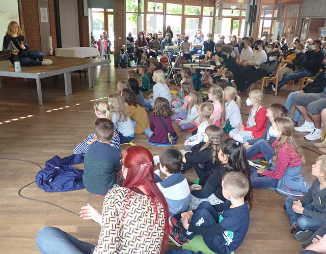 Kinder und Erwachsene hören aufmerksam einer Frau auf der Bühne zu.