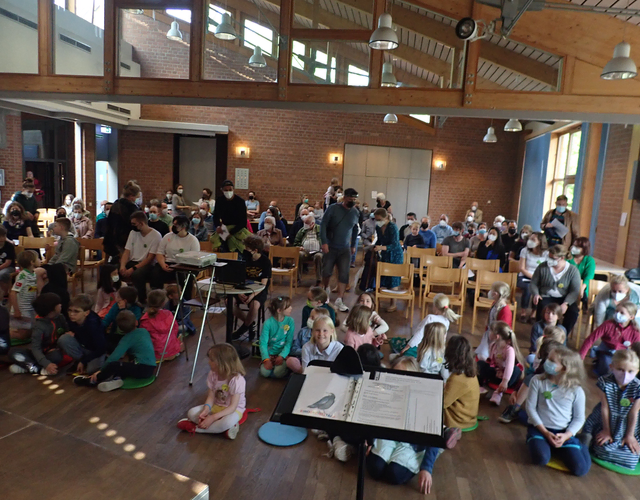 Große Gruppe von Kindern und Erwachsenen sitzt drinnen bei einer Veranstaltung.