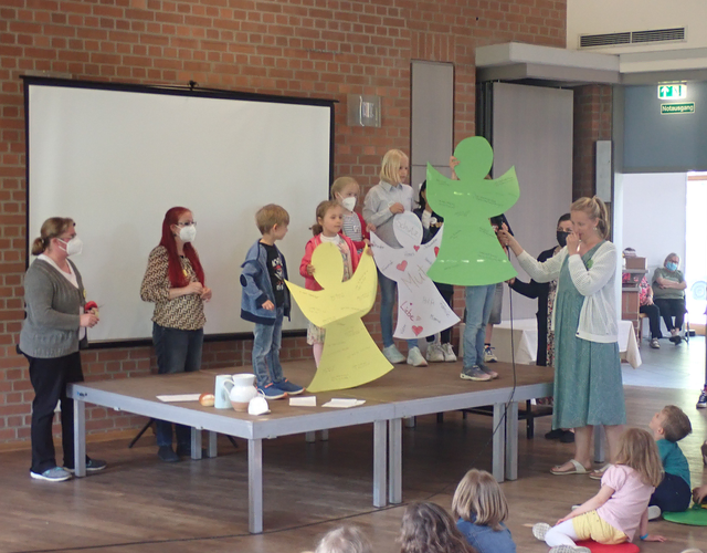 Kinder auf Bühne zeigen bunte Papierengel, Erwachsene helfen, Publikum schaut zu.