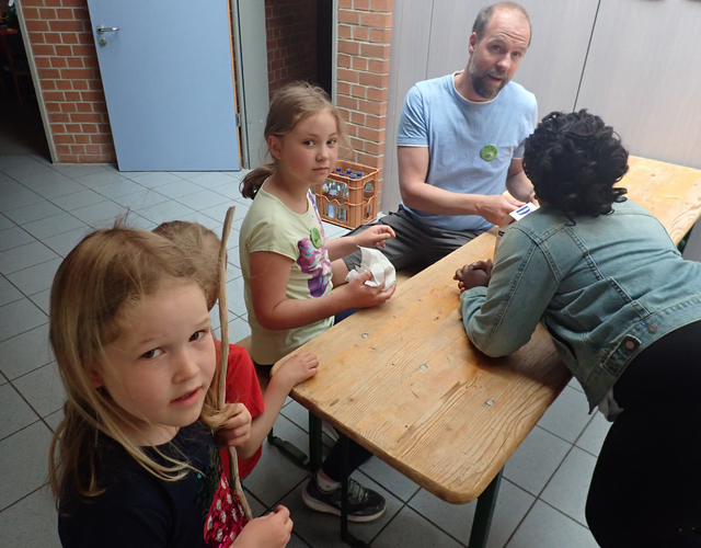 Kinder an Holztisch mit Mann, der drinnen Dinge verteilt.