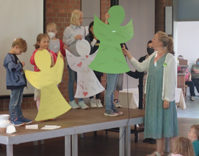 Kinder zeigen große Engelplakate mit Botschaften, Erwachsene spricht mit Mikrofon.