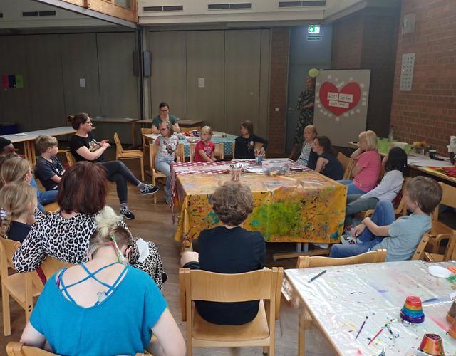Gruppe von Kindern und Erwachsenen im Bastelraum, hört aufmerksam zu.