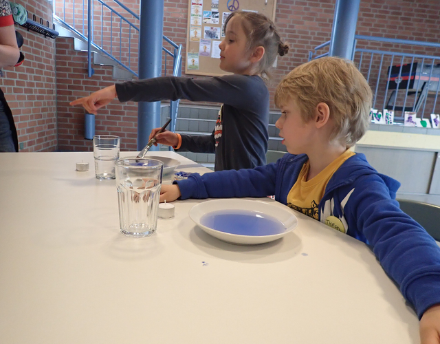 Zwei Kinder am Tisch mit blauem Experiment, eines zeigt neugierig.