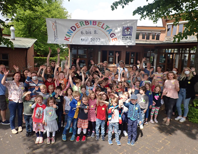Kinder und Erwachsene jubeln draußen unter Kinderbibeltage-Banner, fröhlich lachend.