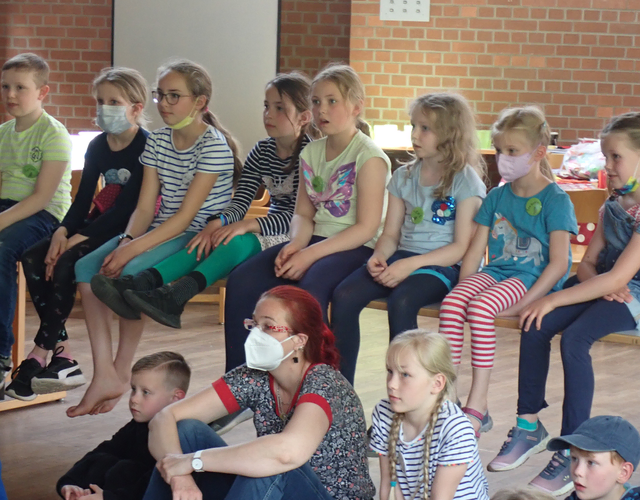 Kinder sitzen drinnen dicht beieinander, schauen aufmerksam, einige mit Masken.