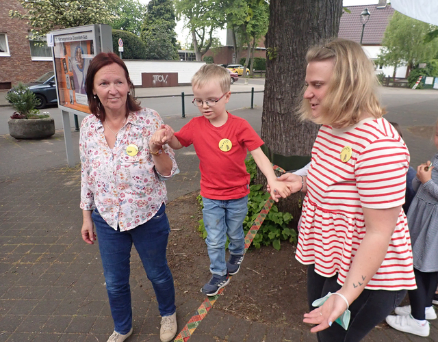 Junge balanciert auf Slackline, wird von zwei Frauen gehalten.