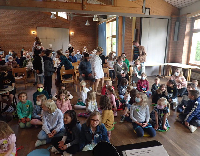 Kinder und Erwachsene sitzen drinnen bei einer gemeinsamen Veranstaltung oder Präsentation.