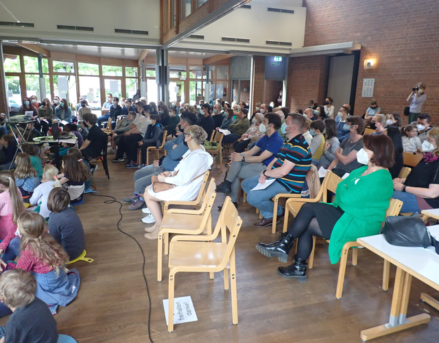 Erwachsene und Kinder verfolgen aufmerksam eine Veranstaltung in einem hellen Saal.