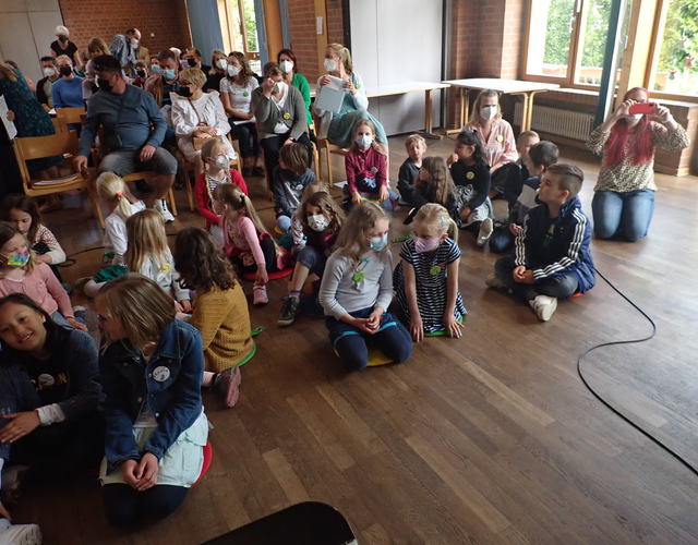 Kinder sitzen vor Bühne, Erwachsene dahinter bei sonniger Veranstaltung drinnen.