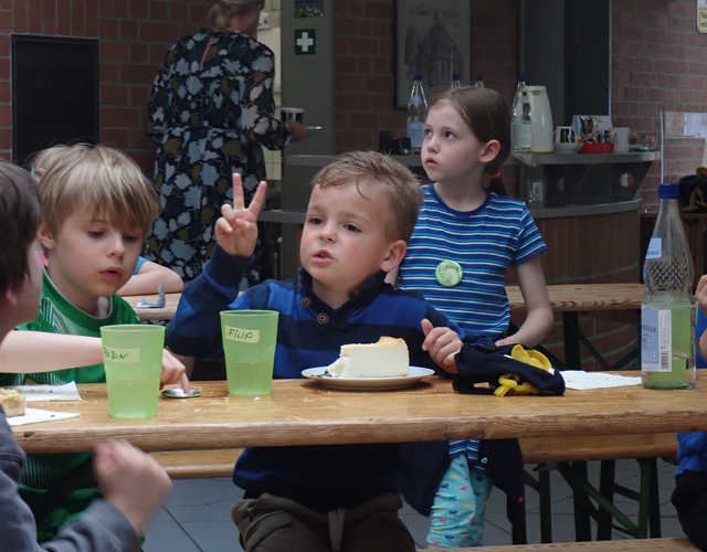 Kinder essen Kuchen am Tisch, eines zeigt das Peace-Zeichen.