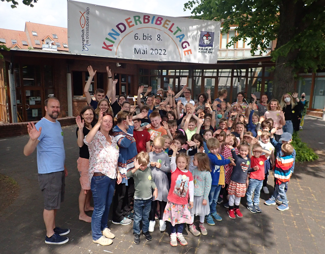 Gruppe winkt fröhlich unter Kinderbibeltage-Banner beim Außenevent, Mai 2022.