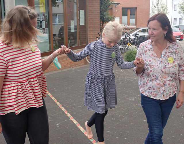 Mädchen balanciert barfuß auf Slackline, von zwei Frauen gehalten.